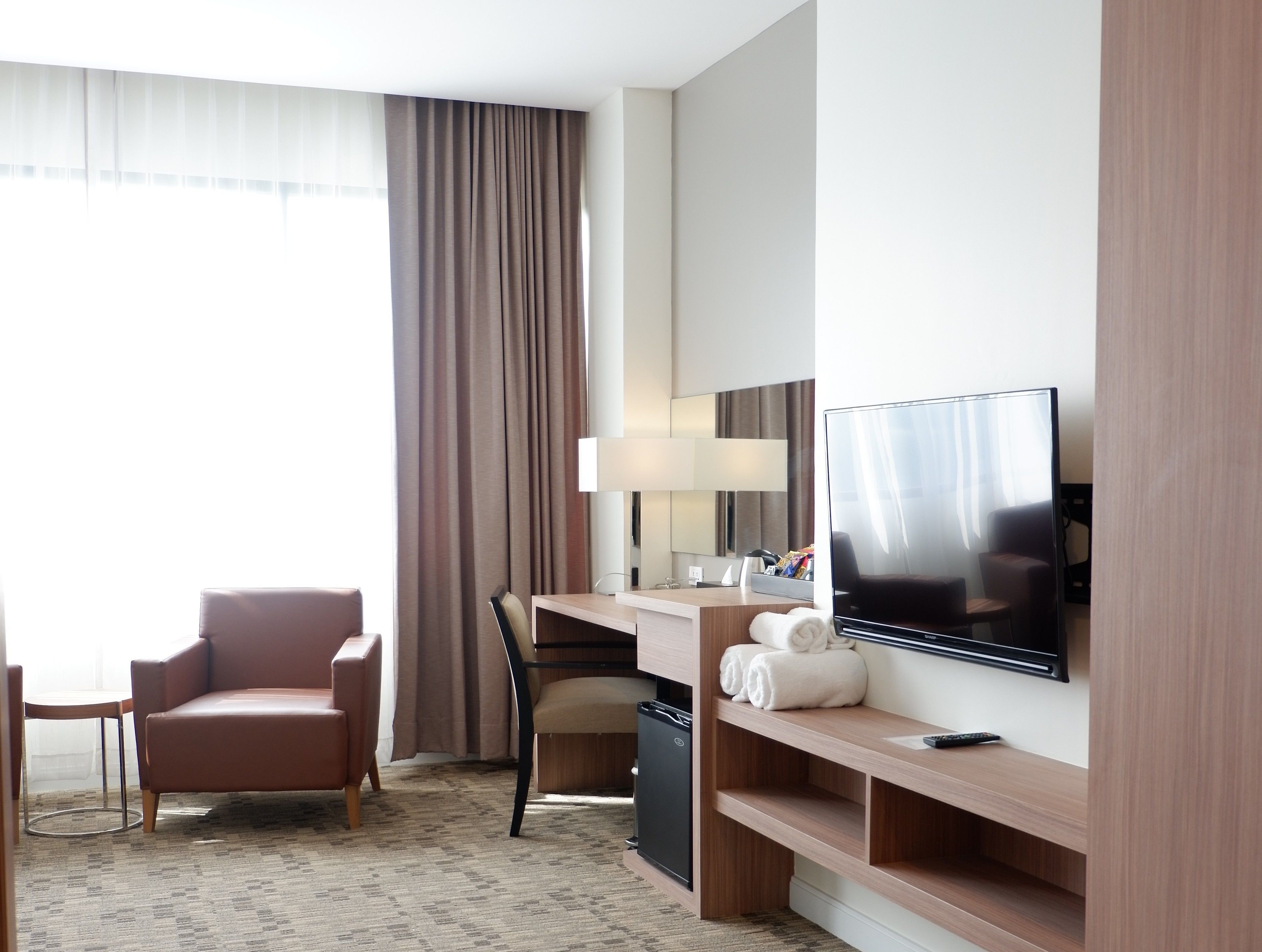 A hotel room with a large flat-screen TV, a desk with a lamp, a chair, and a sofa.
