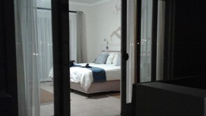 A bedroom with a bed, a nightstand, and a window with curtains. - Stay at Swakop Guesthouse (Swakopmund)