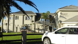 A two-story building with a sign for 'SWAKOP' and a contact number. There are palm trees and outdoor seating areas visible. - Stay at Swakop Guesthouse (Swakopmund)