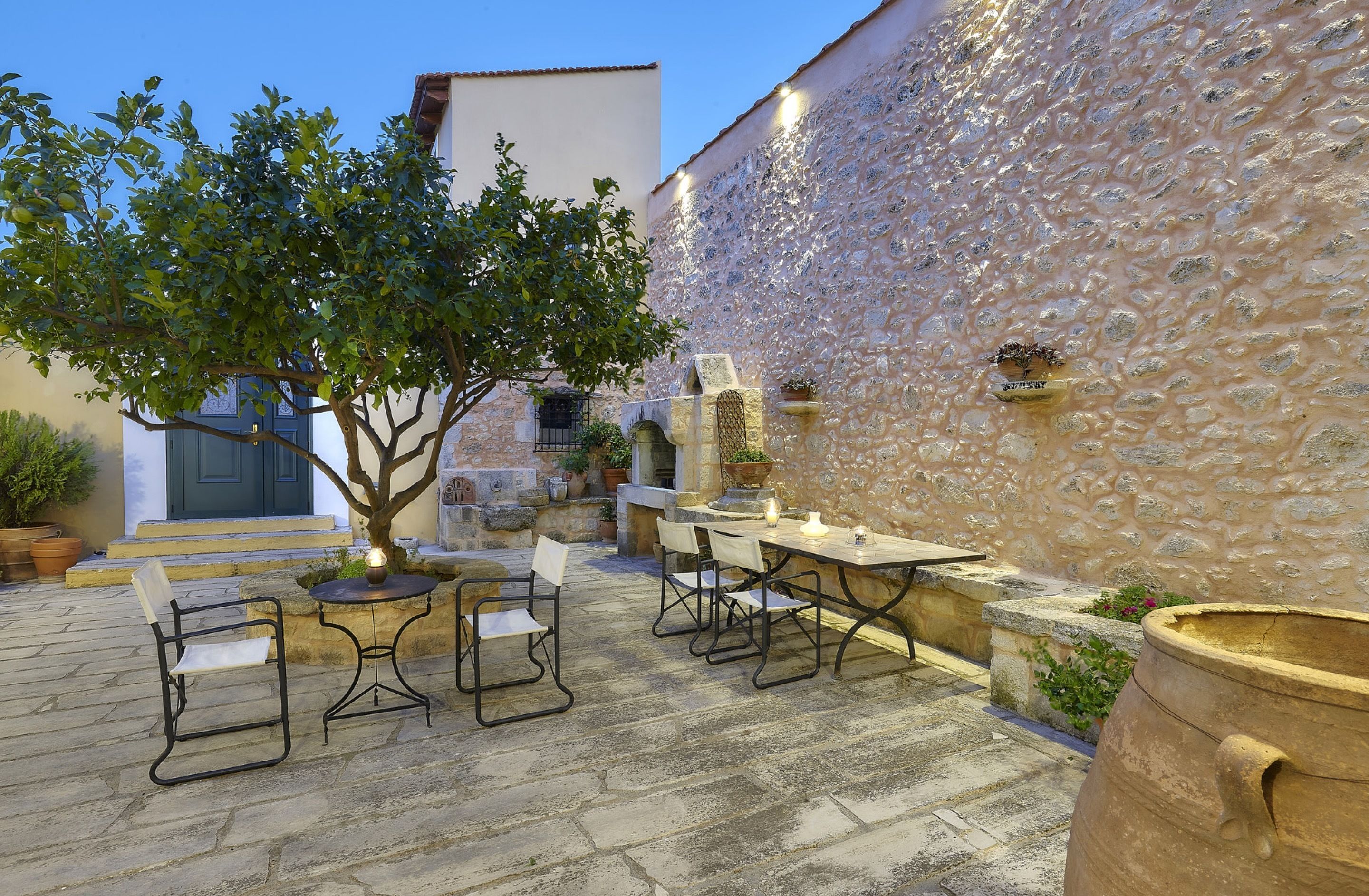 Une terrasse extérieure avec un sol en pierre, une table avec des chaises, un arbre et un mur en pierre.