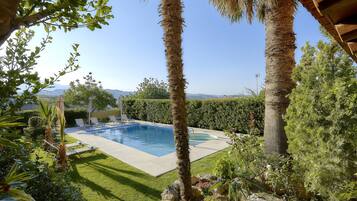 Relax by the serene pool surrounded by lush greenery and palm trees, offering a tranquil retreat with a view of distant hills.