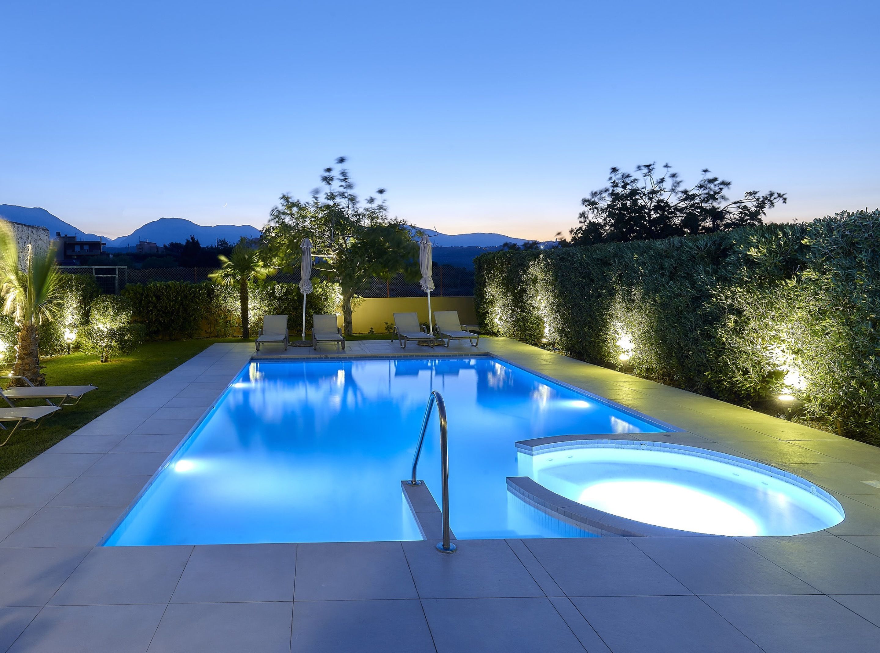 A rectangular swimming pool with a diving board, surrounded by lounge chairs and umbrellas, with a view of mountains in the background.