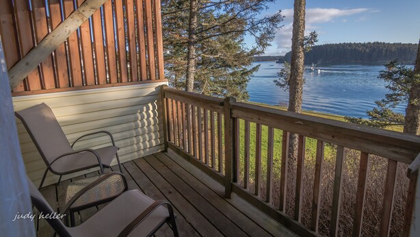 A wooden deck with two chairs and a table, overlooking a lake with trees and a distant shoreline. - Kodiak Awesome View Vacation Rentals: The  Captains Quarters (Kodiak)