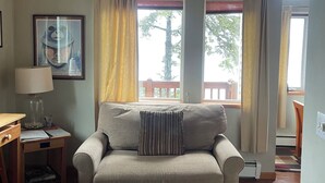 A living room with a sofa, a lamp, a framed picture, and a window with curtains. - Kodiak Awesome View Vacation Rentals: The  Captains Quarters (Kodiak)