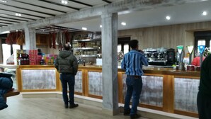A cafe with a wooden counter and a coffee machine. There are people standing at the counter. - Pensión El Carloto (Campoo de Yuso)