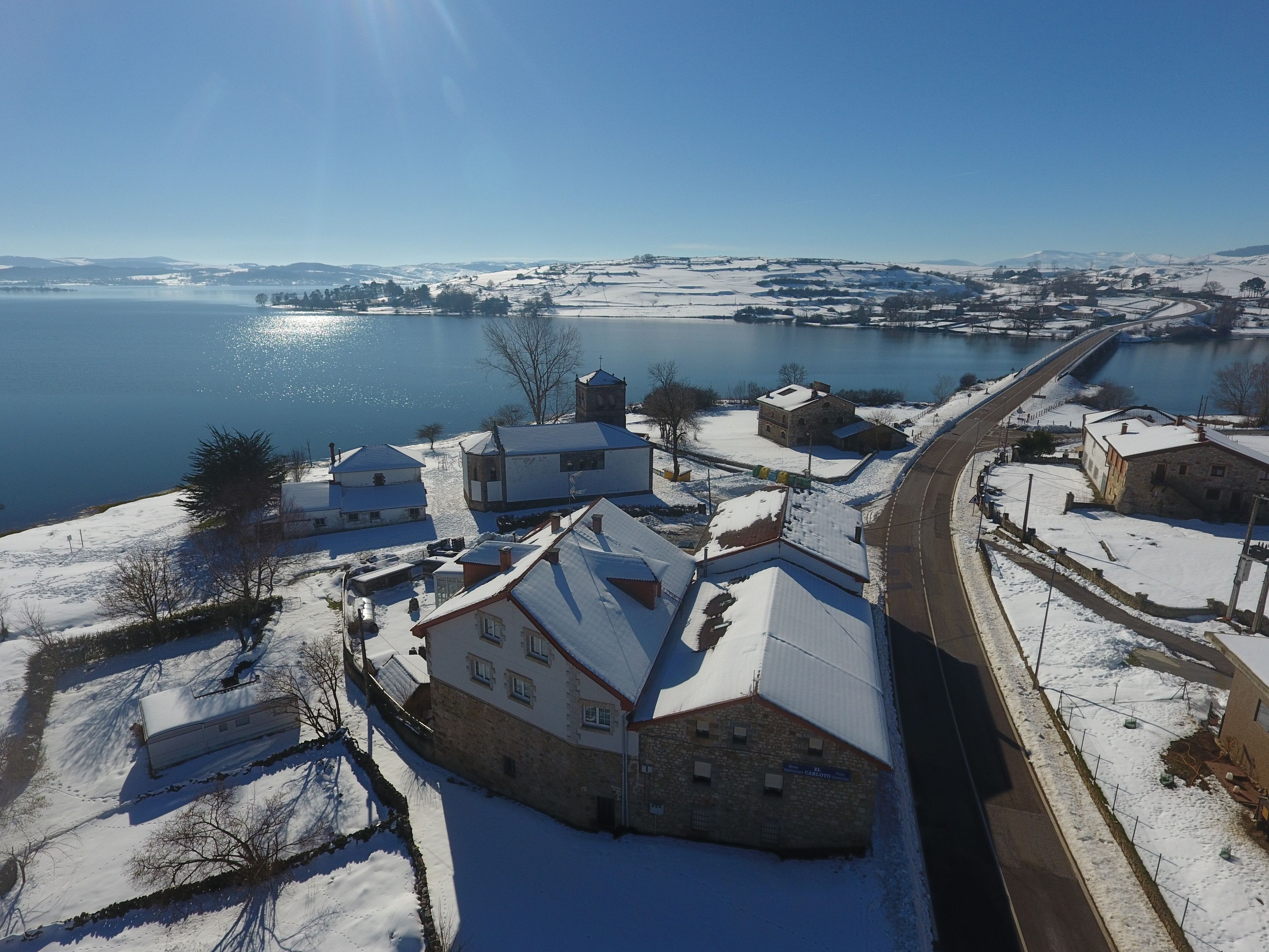 Un pueblo cubierto de nieve con edificios de piedra tradicionales, una carretera y un lago al fondo.