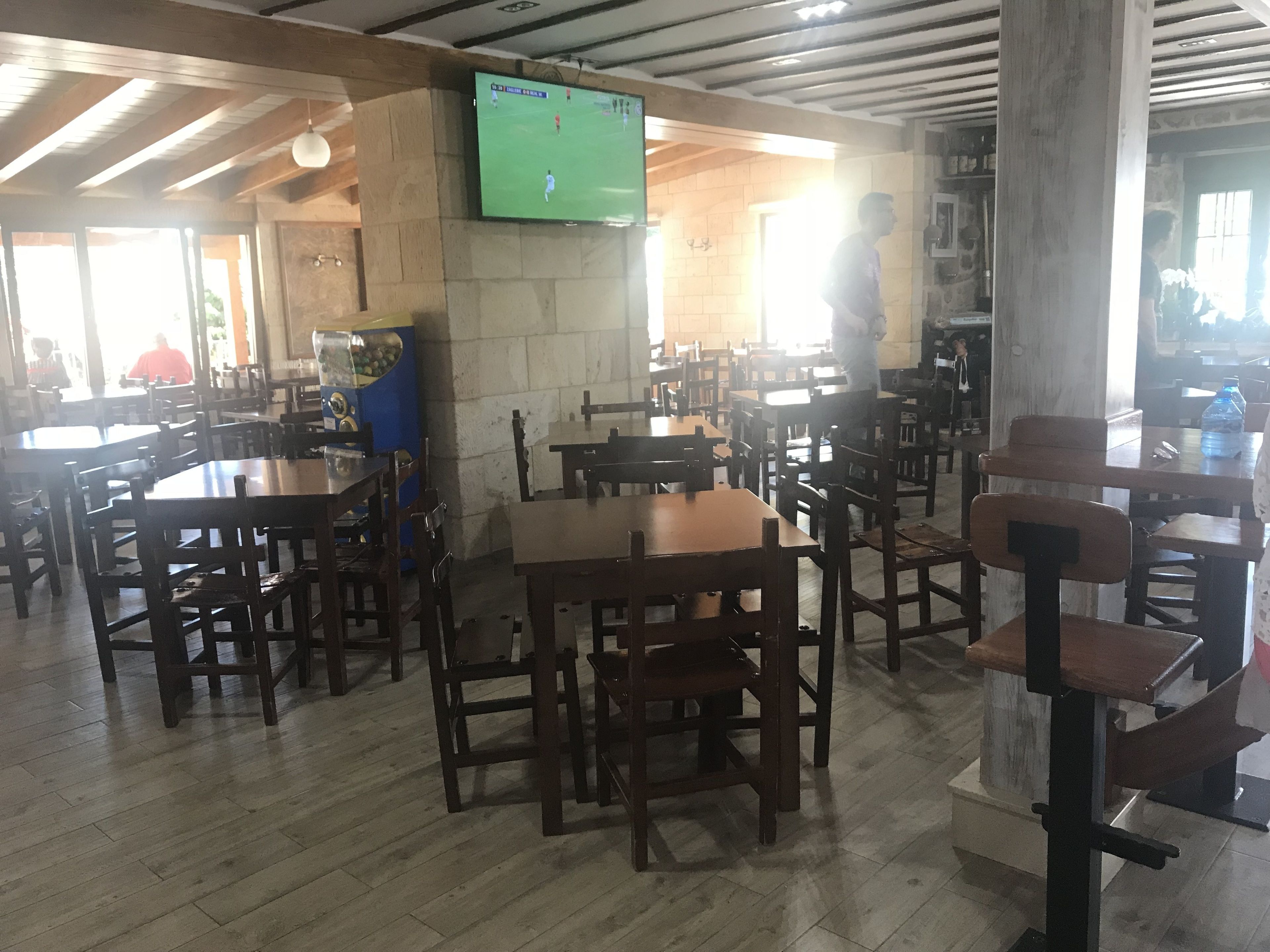 Un restaurante con mesas y sillas de madera, un televisor montado en la pared y una máquina expendedora.