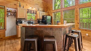 A wooden cabin interior with a kitchen, bar stools, and large windows.