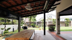 An outdoor patio with wooden tables and benches, a ceiling fan, and a view of a garden with various plants and a barbecue grill.