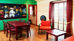 Una habitación con suelo de madera, una mesa de comedor con sillas, un sillón rojo, una ventana con cortinas rojas y una pared con un mural colorido.