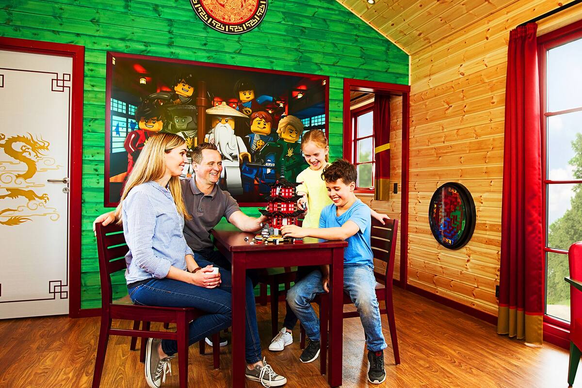 Uma família brincando com LEGO em uma mesa em um quarto com paredes de madeira e uma janela grande.