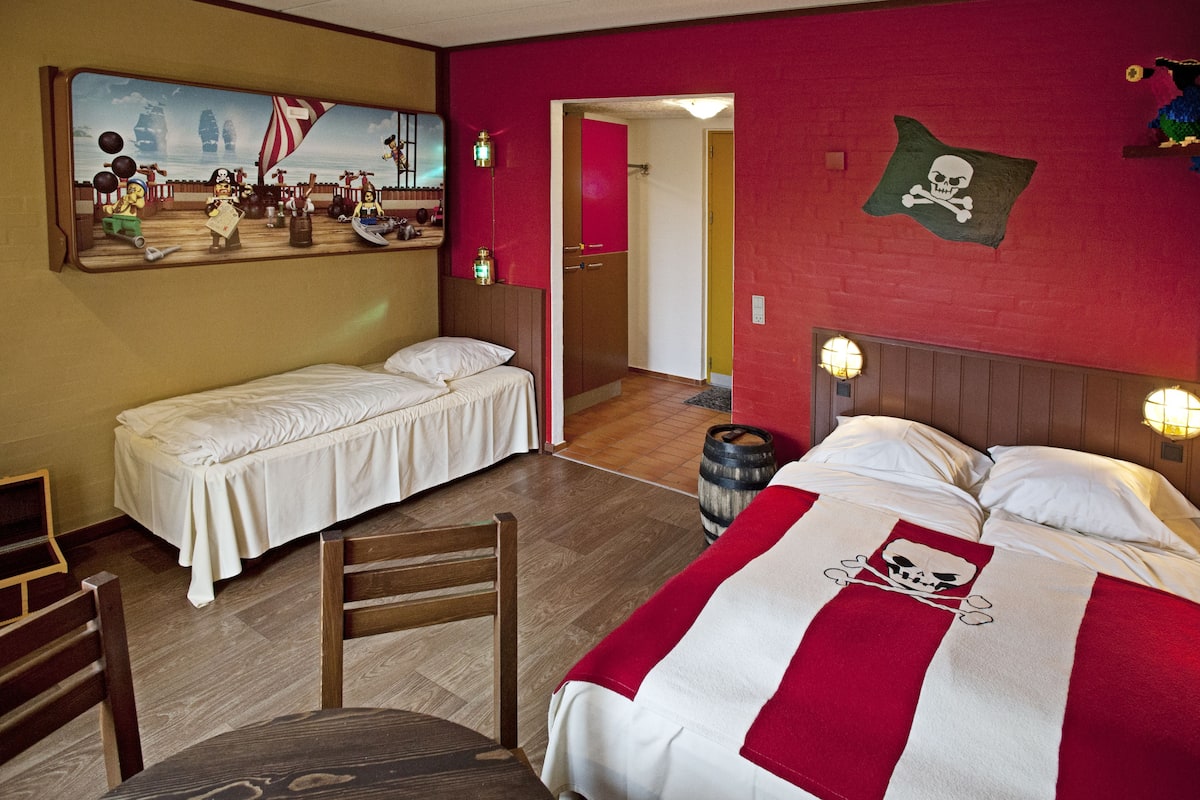 A room with two beds, a wooden chair, a table, and a painting on the wall.