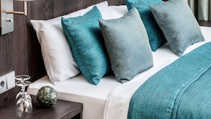 A neatly made bed with teal and white bedding, a bedside lamp, and a small table with a glass and a decorative item. - Hotel Concordia (Euskirchen)