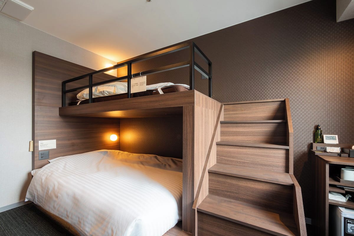 Habitación de hotel compacta con una litera, una escalera de madera, un escritorio con un reloj y una lavadora Procter & Gamble.