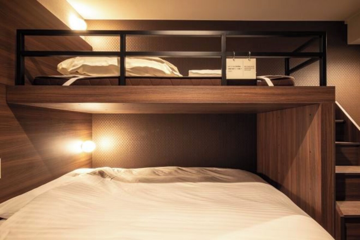 A bunk bed with a wooden frame and a mattress. There is a small table with a chair and a light fixture on the wall.