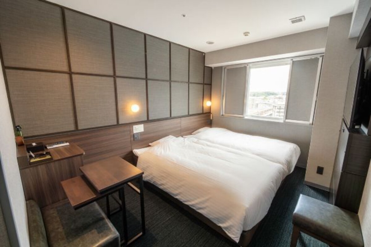 A modern hotel room with a large bed, a small desk, a chair, and a window with a city view.