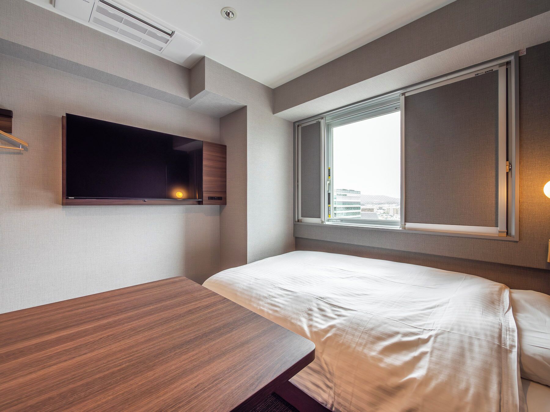 A modern hotel room with a bed, a wooden desk, a flat-screen TV mounted on the wall, and a window with a city view.