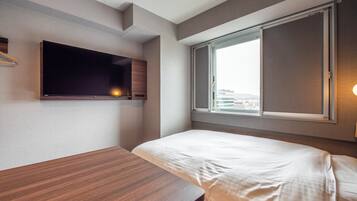 A modern hotel room with a bed, a wooden desk, a flat-screen TV mounted on the wall, and a window with a city view.