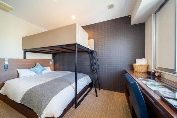 Une chambre d’hôtel avec un lit superposé, un bureau avec une chaise, une fenêtre et un téléphone fixé au mur.