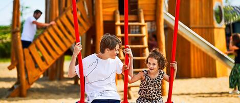 A wooden play structure with swings and a slide, surrounded by sand.