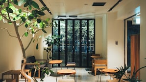 Un interior moderno con una zona de estar, mesas y sillas de madera, rodeado de plantas en macetas.