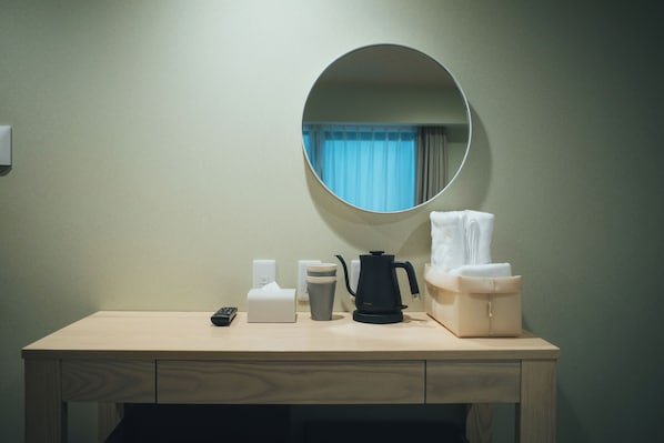 Habitación de hotel con un escritorio de madera, un espejo redondo, cafetera, hervidor de agua, caja de pañuelos, taza y cesto para toallas.