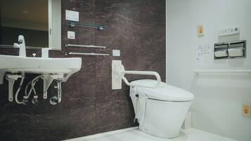 A modern bathroom with a marble wall, a white sink, a toilet with a grab bar, and a hand sanitizer dispenser.