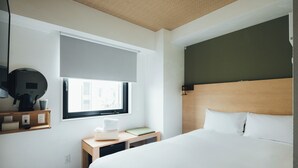 Habitación de hotel con una cama grande, una mesita de noche con lámpara, un escritorio pequeño con silla, una ventana con persiana y un televisor montado en la pared.