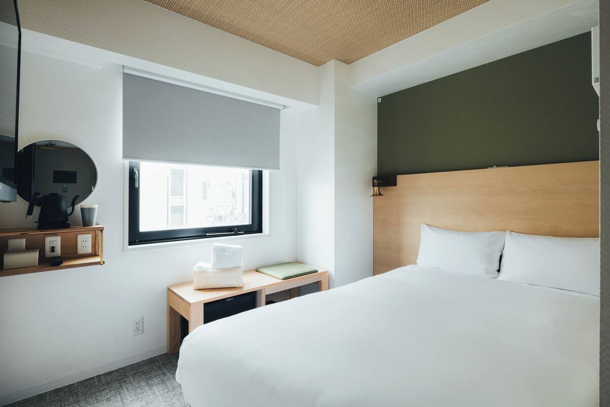 Hotellihuone, jossa on suuri sänky, yöpöytä lampun kanssa, pieni työpöytä tuolilla, ikkuna, jossa on sälekaihtimet, ja seinälle asennettu televisio.