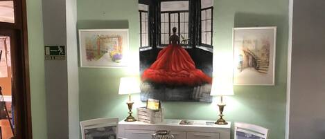 A room with a green wall, a painting of a woman in a red dress, two chairs, a small table, and a chandelier.