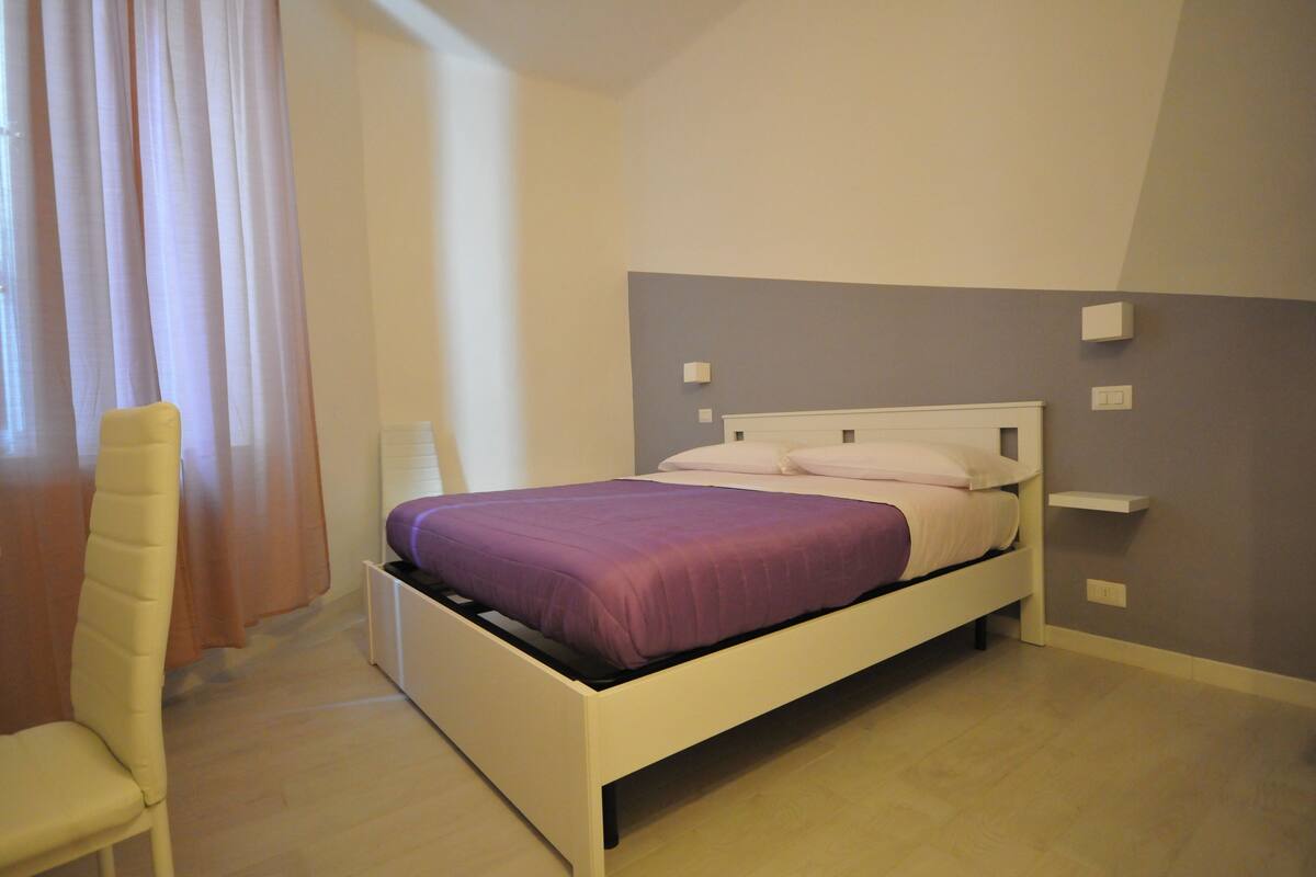 A bedroom with a white bed, a chair, and a window with curtains.