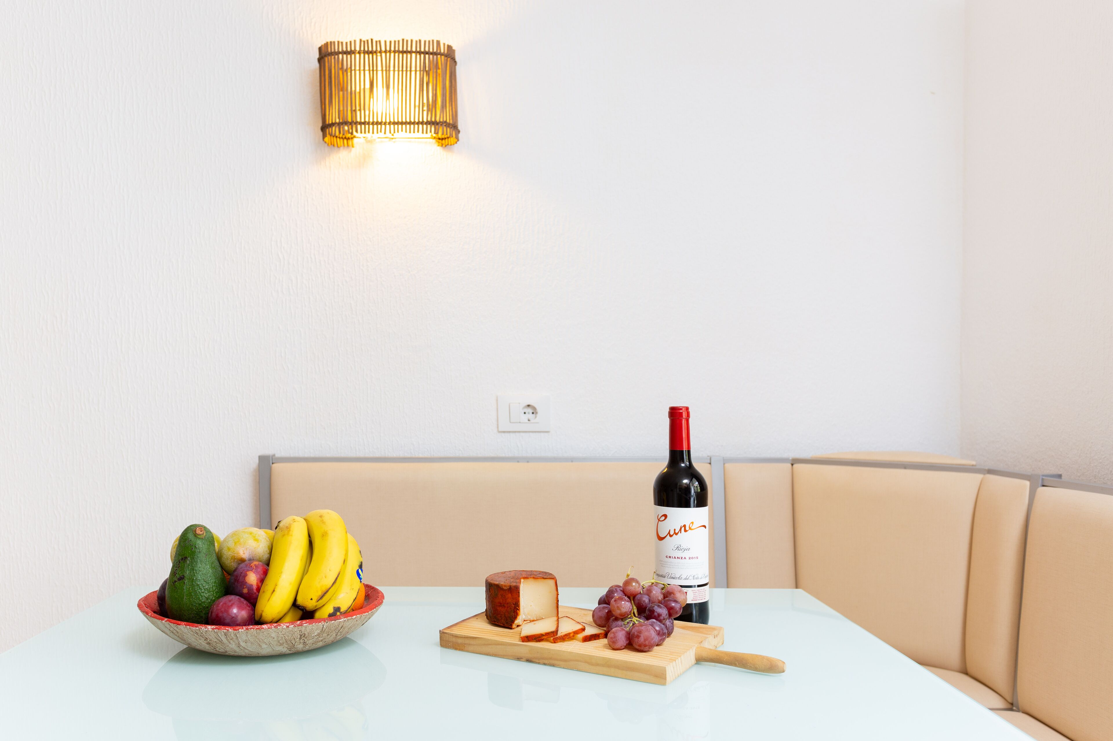A dining table with a bowl of fruit, a cheese wedge, a bottle of wine, and a cutting board with grapes.