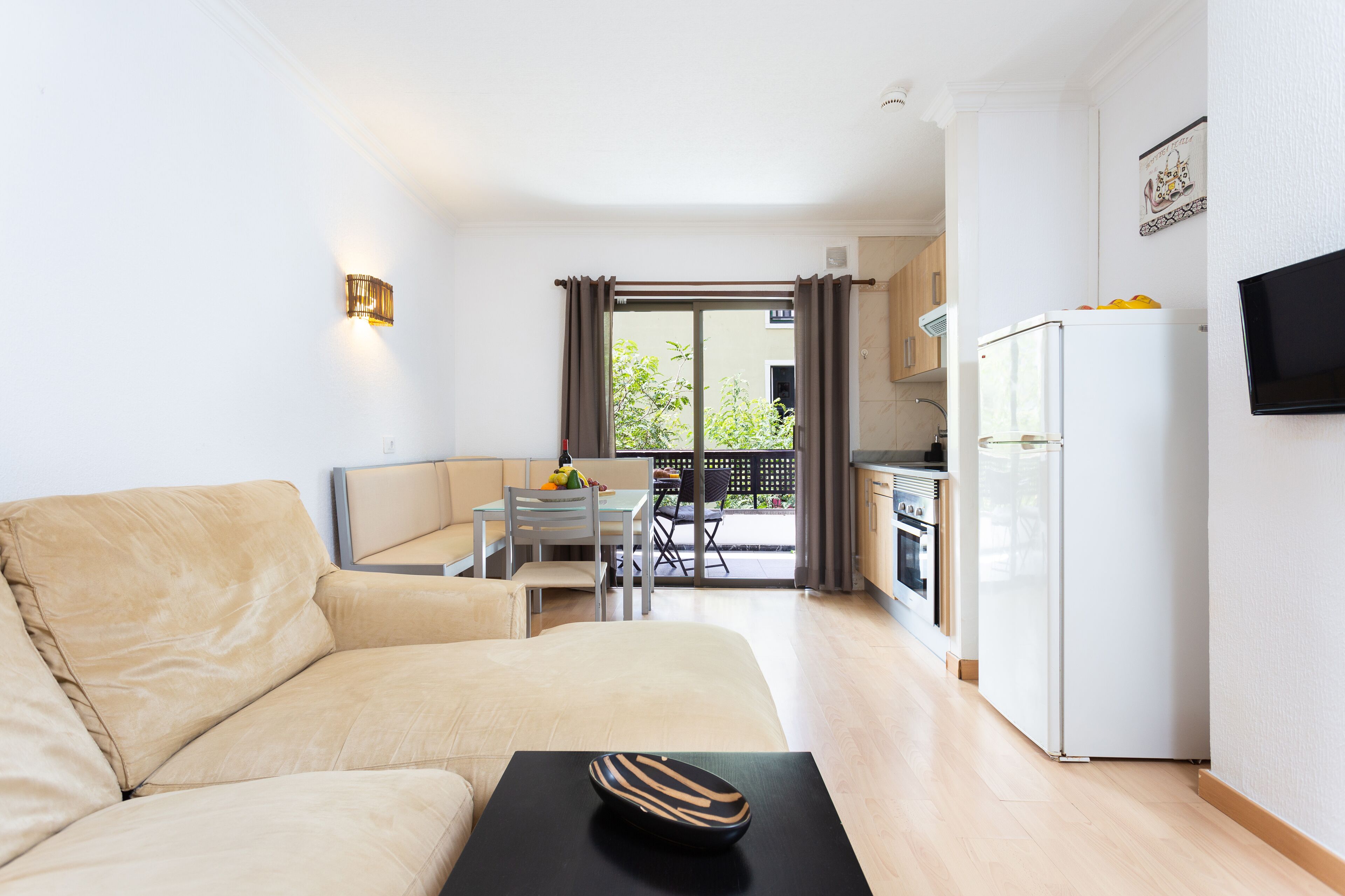 Una sala moderna con sofá, una mesa de comedor con sillas, una zona de cocina con refrigerador y un televisor montado en la pared.