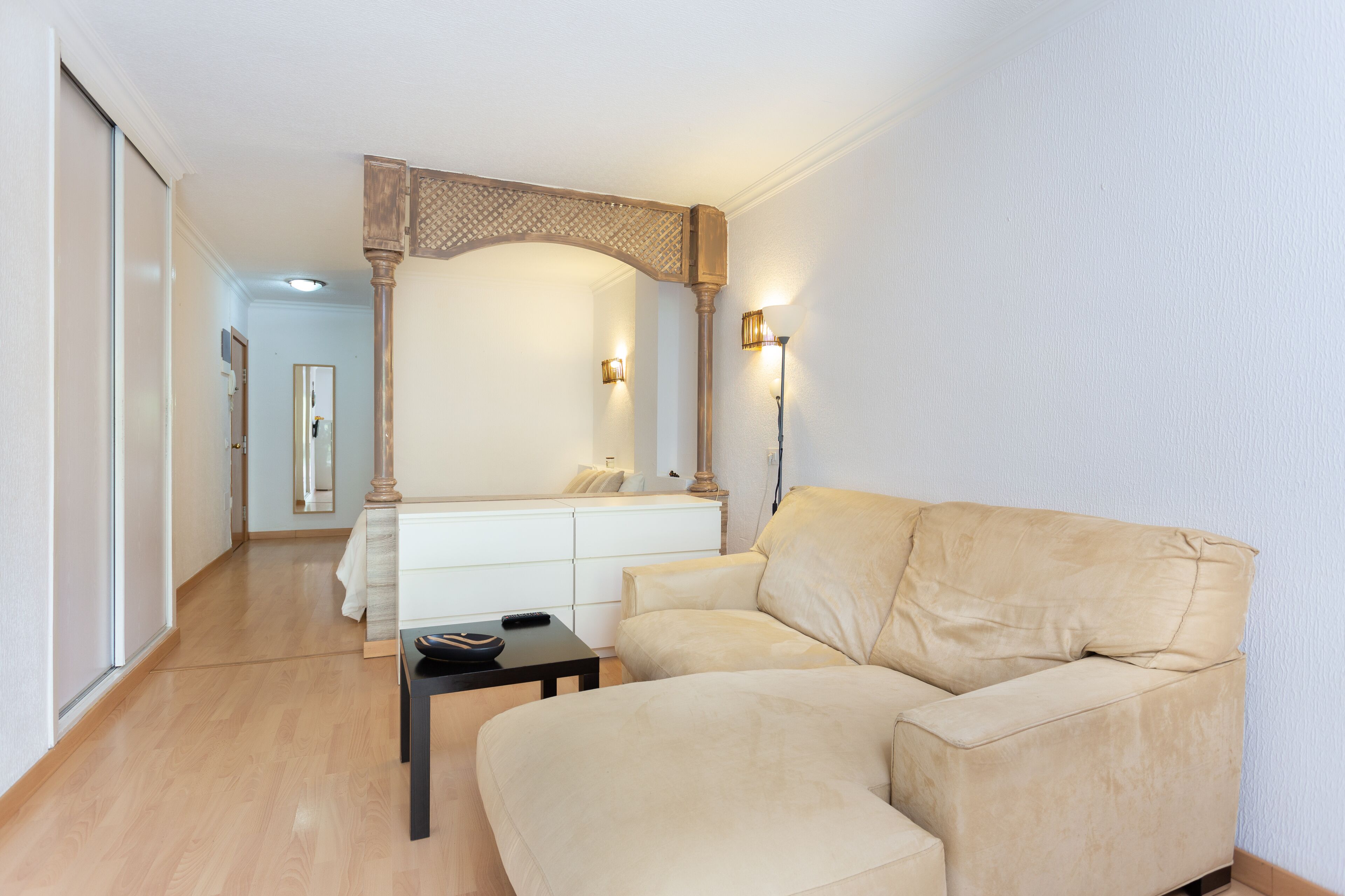 Una sala de estar moderna con un sofá color beige, una mesita negra pequeña y una consola blanca. Se aprecia una viga de techo de madera y dos luces fijadas a la pared.