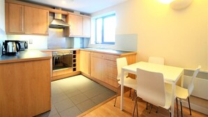 A compact kitchen with wooden cabinets, a built-in oven, and a dining area with a white table and chairs. - City Walk (Leeds)