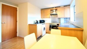 A compact kitchen with wooden cabinets, a microwave, a washing machine, and a dining area with a table and chairs. - City Walk (Leeds)