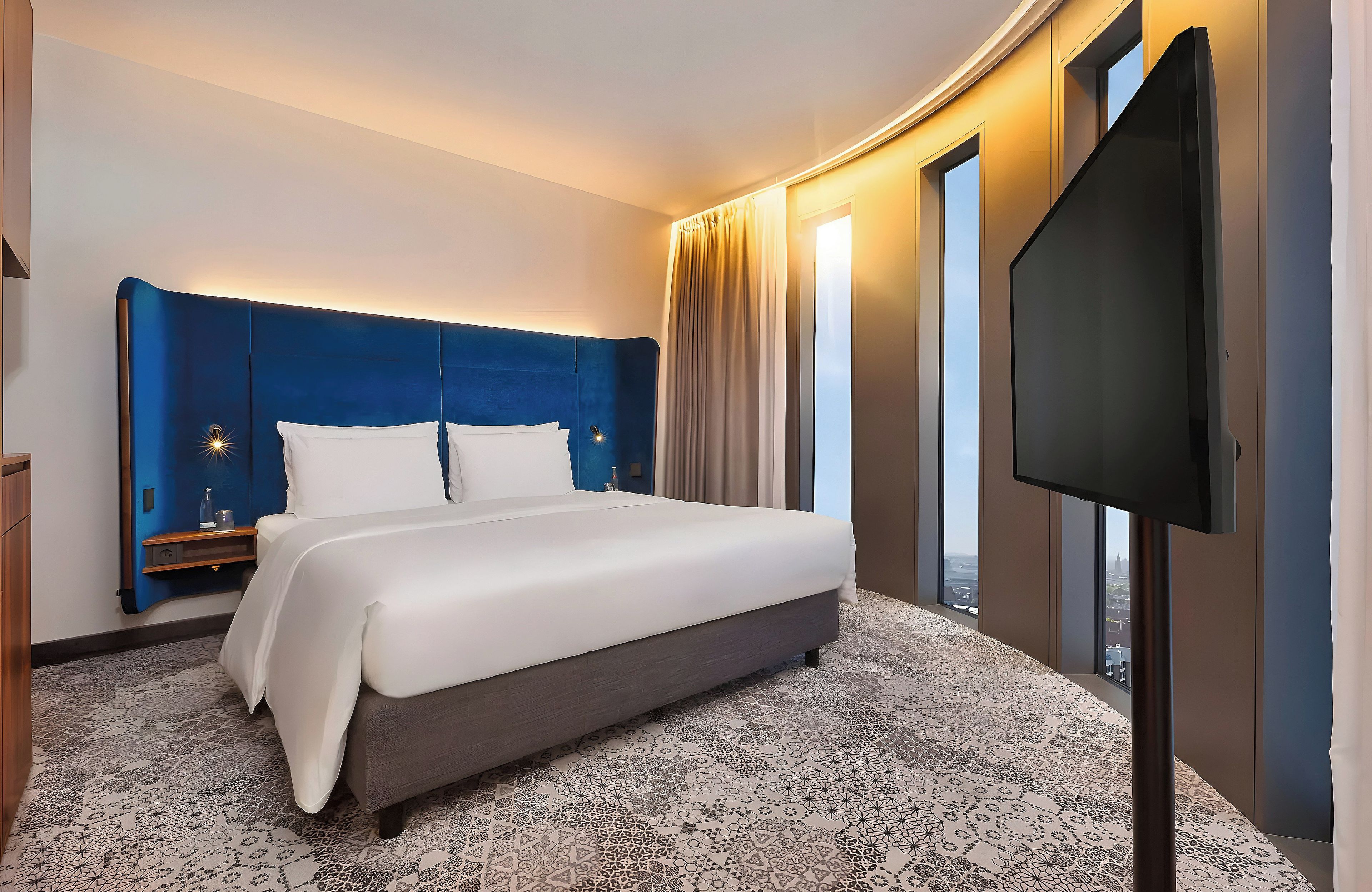 A hotel room with a large bed, a flat-screen TV, a nightstand, and a view of the city through large windows.