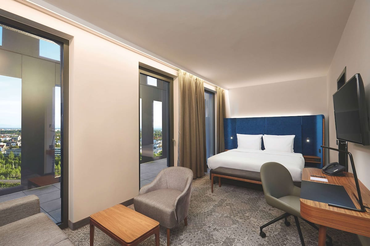 Habitación de hotel con cama, escritorio, silla, televisor y balcón con vistas.