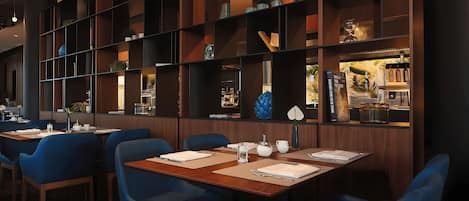 A restaurant interior with multiple tables and chairs, wooden shelves with decorative items, and a bar area with bottles.