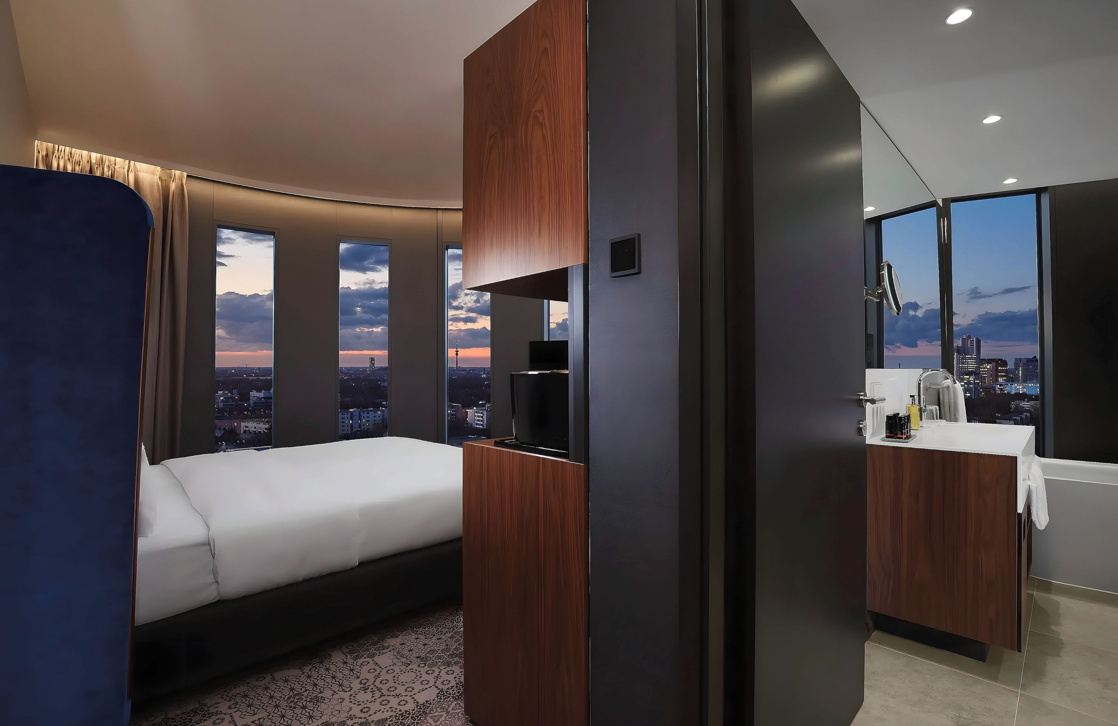 Habitación de hotel moderna con cama, televisor, baño con lavamanos y ventanas amplias con vista a la ciudad.