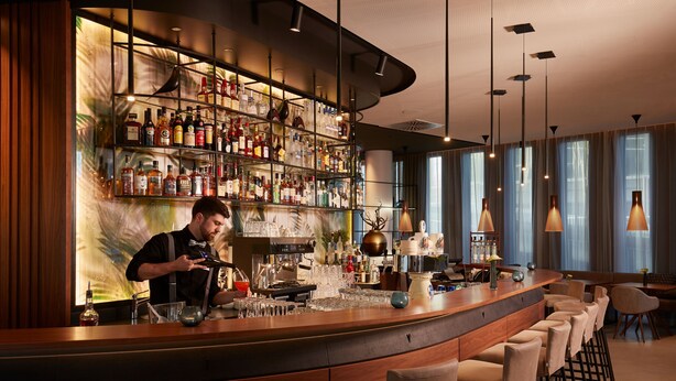 A bar area with a bartender, a well-stocked liquor shelf, and a seating arrangement with bar stools. - HYPERION Hotel München (Munich)