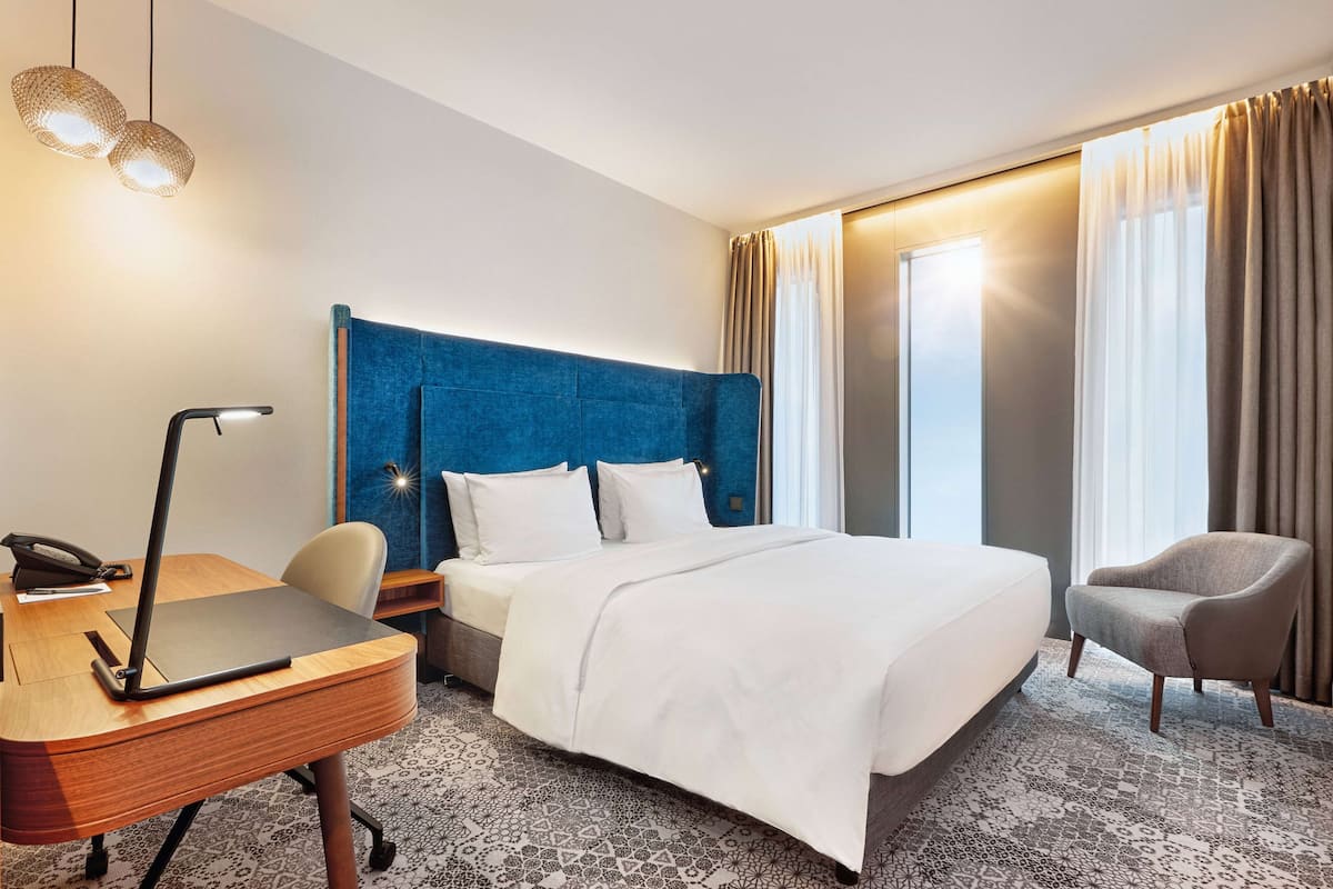 Habitación de hotel con una cama grande, un escritorio con lámpara, una silla y una ventana con cortinas.