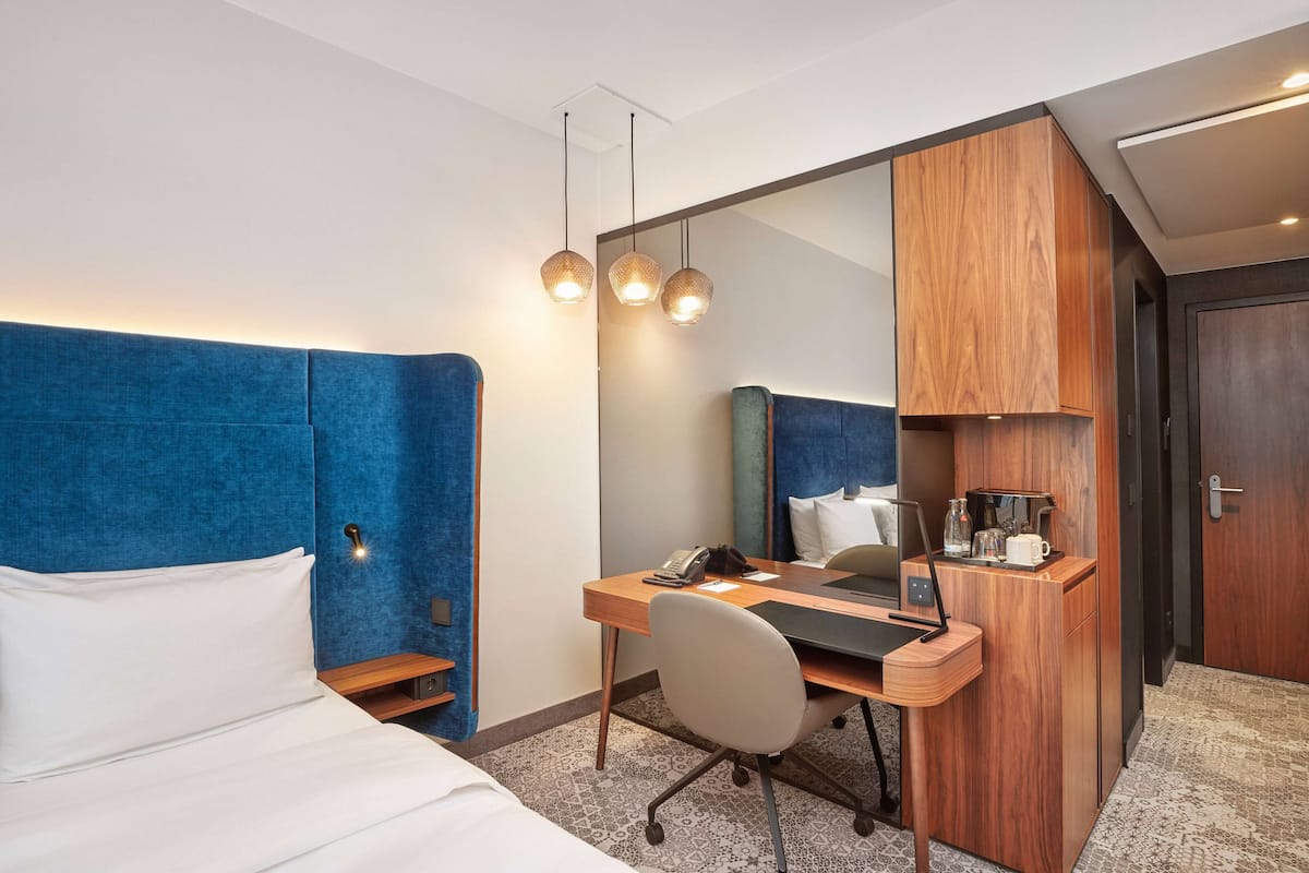 Habitación de hotel con cama, escritorio con laptop, silla, cafetera y un espejo grande.