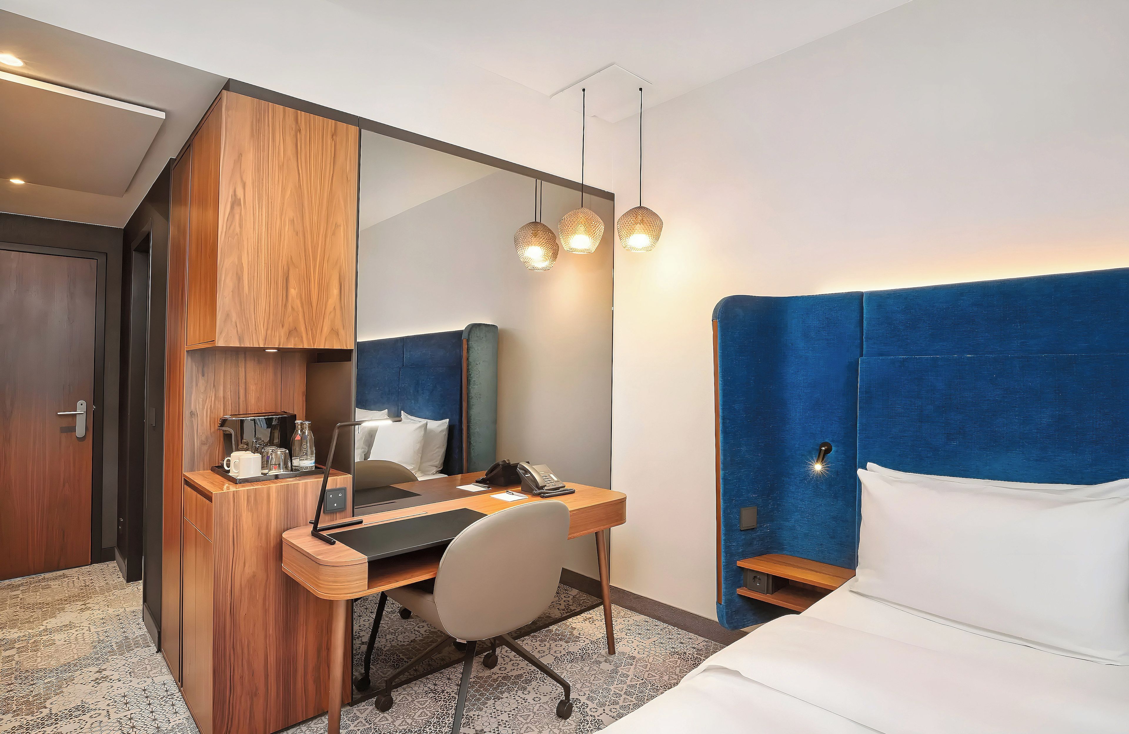 Ein Hotelzimmer mit einem Holztisch, einem Bett mit blauer Kopfteil, einem großen Spiegel und einer Hängelampe.