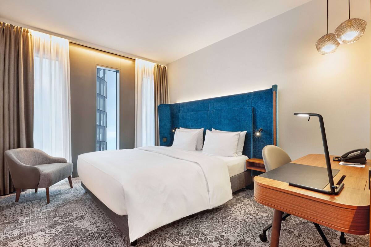 Habitación de hotel con una cama grande, un escritorio con lámpara, una silla y vistas a la ciudad desde la ventana.