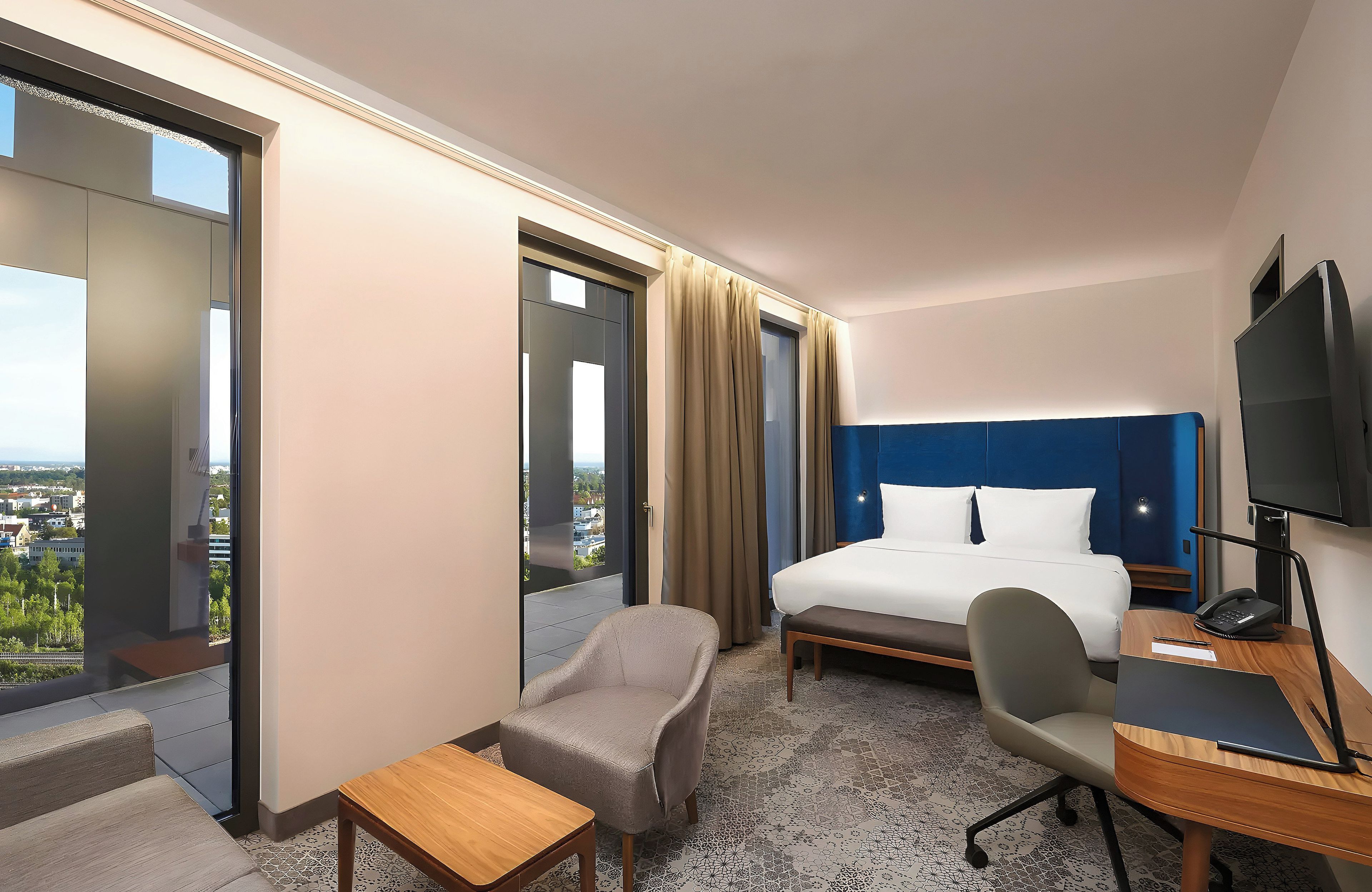 A hotel room with a bed, a desk, a chair, a TV, and a view of the city.