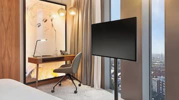 A hotel room with a desk, chair, television, and a large window with a city view.