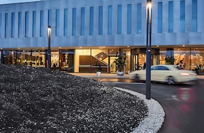 A modern building with a glass facade, a landscaped area with rocks, and a street with cars.