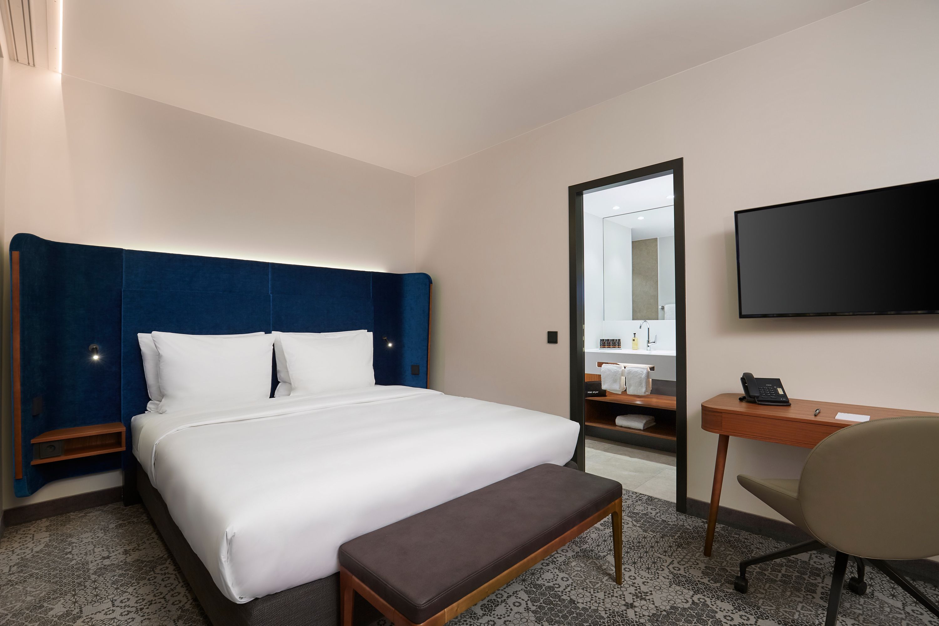 A hotel room with a large bed, a desk, a chair, a flat-screen TV, and a bathroom visible through a mirror.