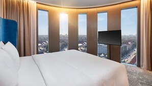 A hotel room with a large bed, a flat-screen TV, and a city view through large windows. - HYPERION Hotel München (Munich)
